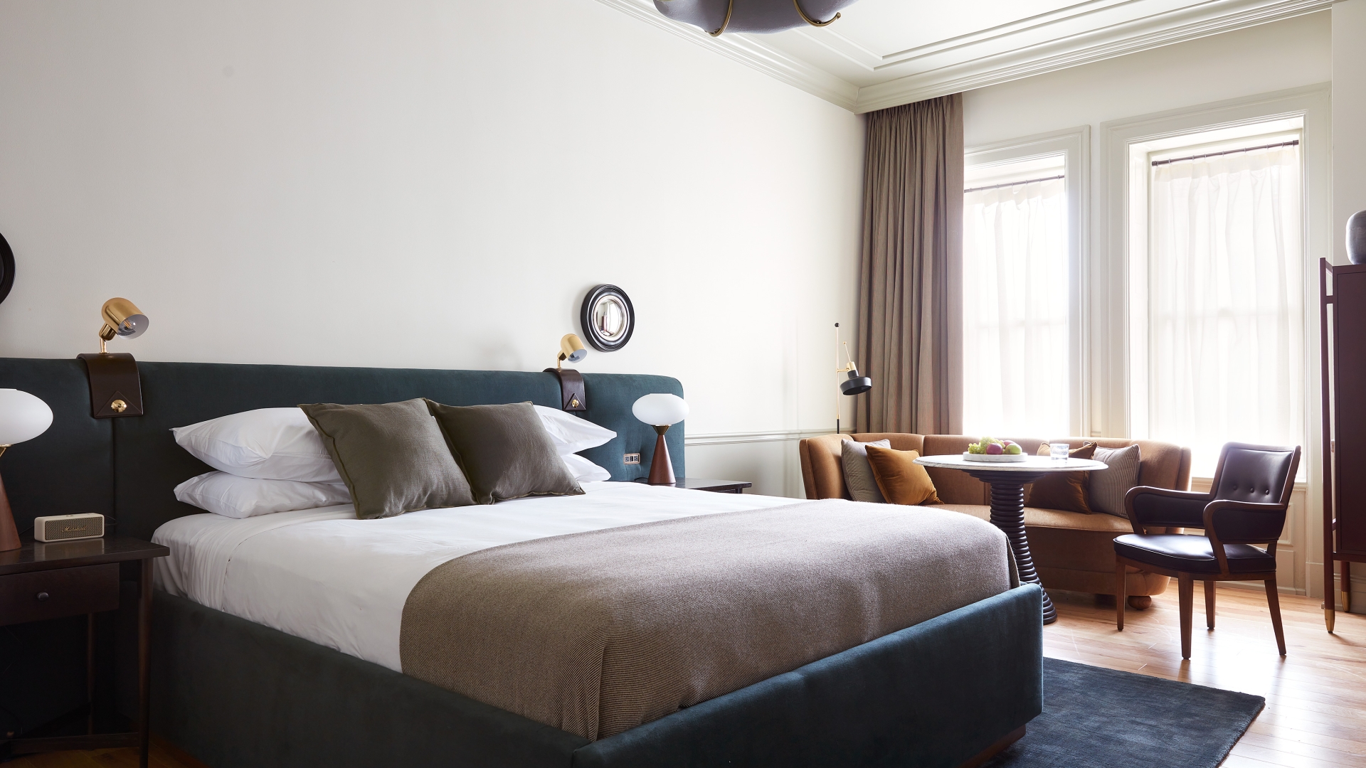 A wide shot of a guest room with an upholstered velvet king size bed and a sitting area with a sofa, table and chair. There are tall, sunny windows, hardwood floors, and a carpet under the bed.