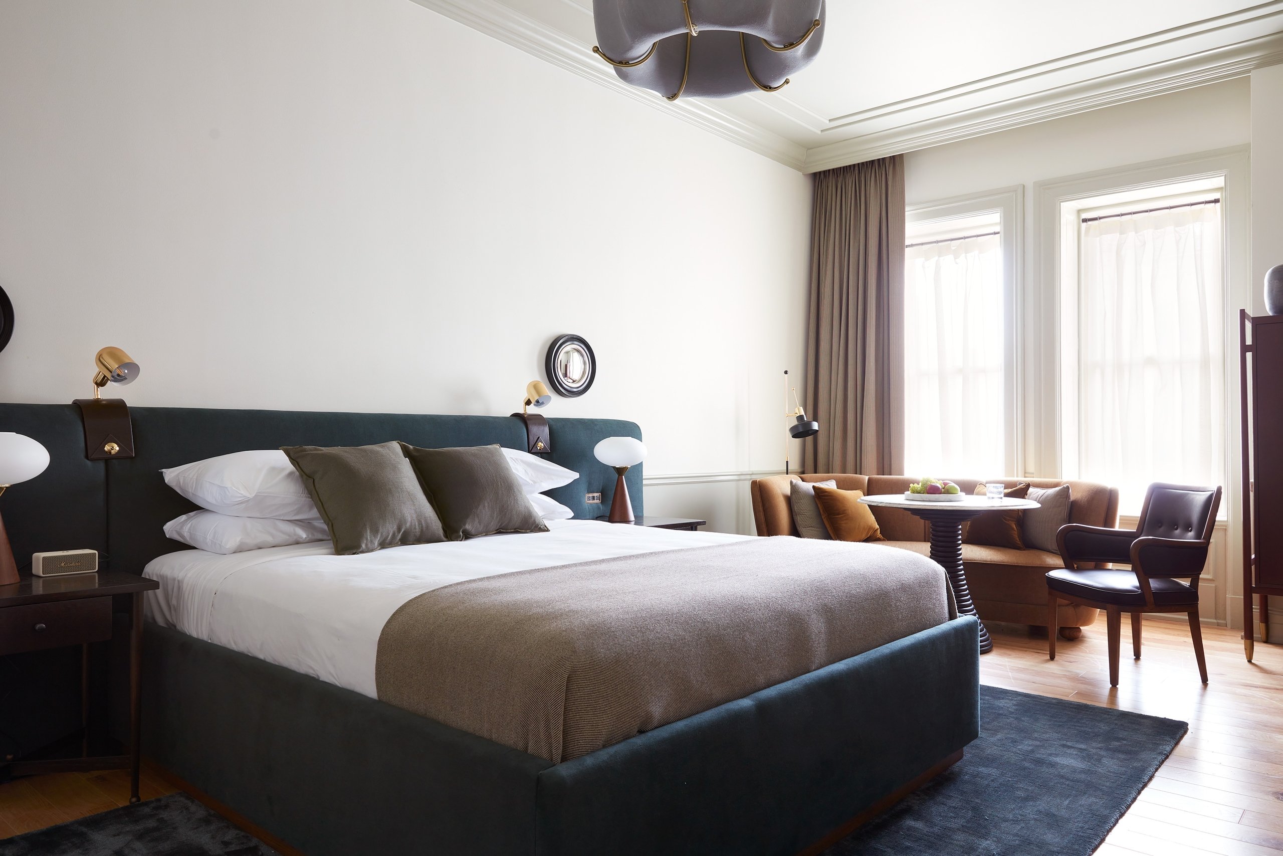 A wide shot of a guest room with an upholstered velvet king size bed and a sitting area with a sofa, table and chair. There are tall, sunny windows, hardwood floors, and a carpet under the bed.