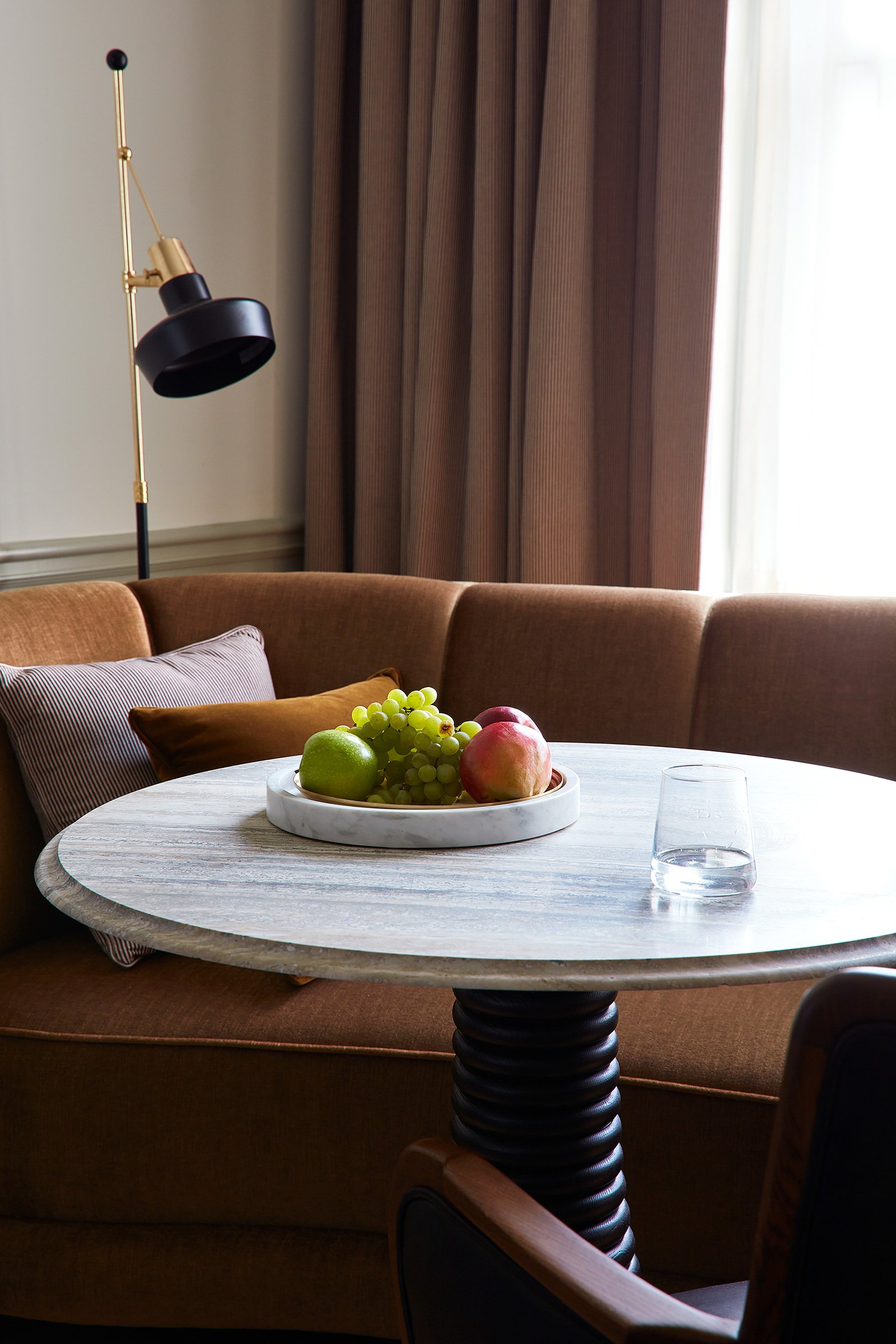 A white stone coffee table with a bowl of fruit on top, in front of a brown couch by the window.