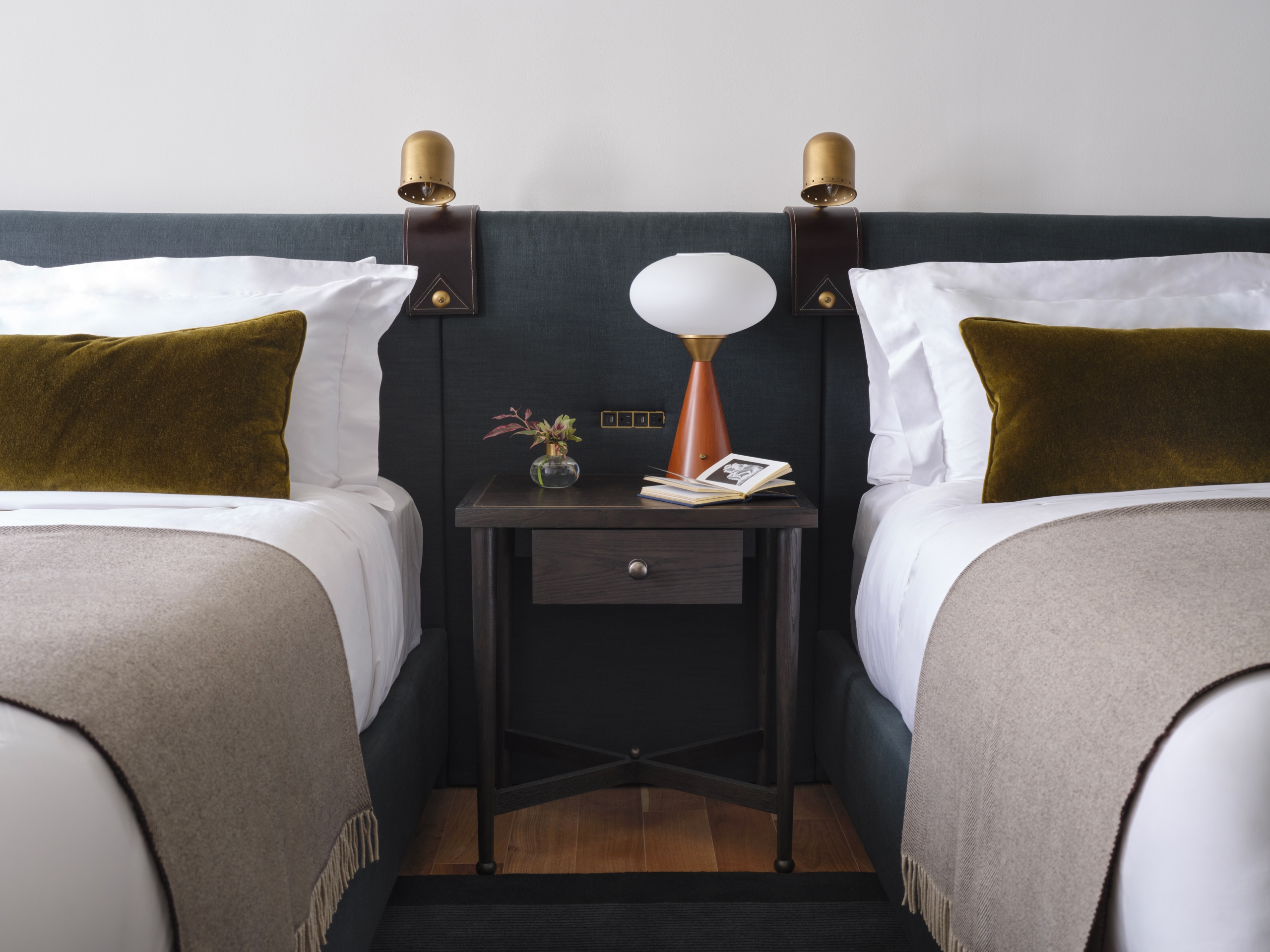 A closeup of two queen beds against a navy velvet upholstered headboard. There is a slim wood nightstand between with a Mid-Century table lamp and a small floral vase.