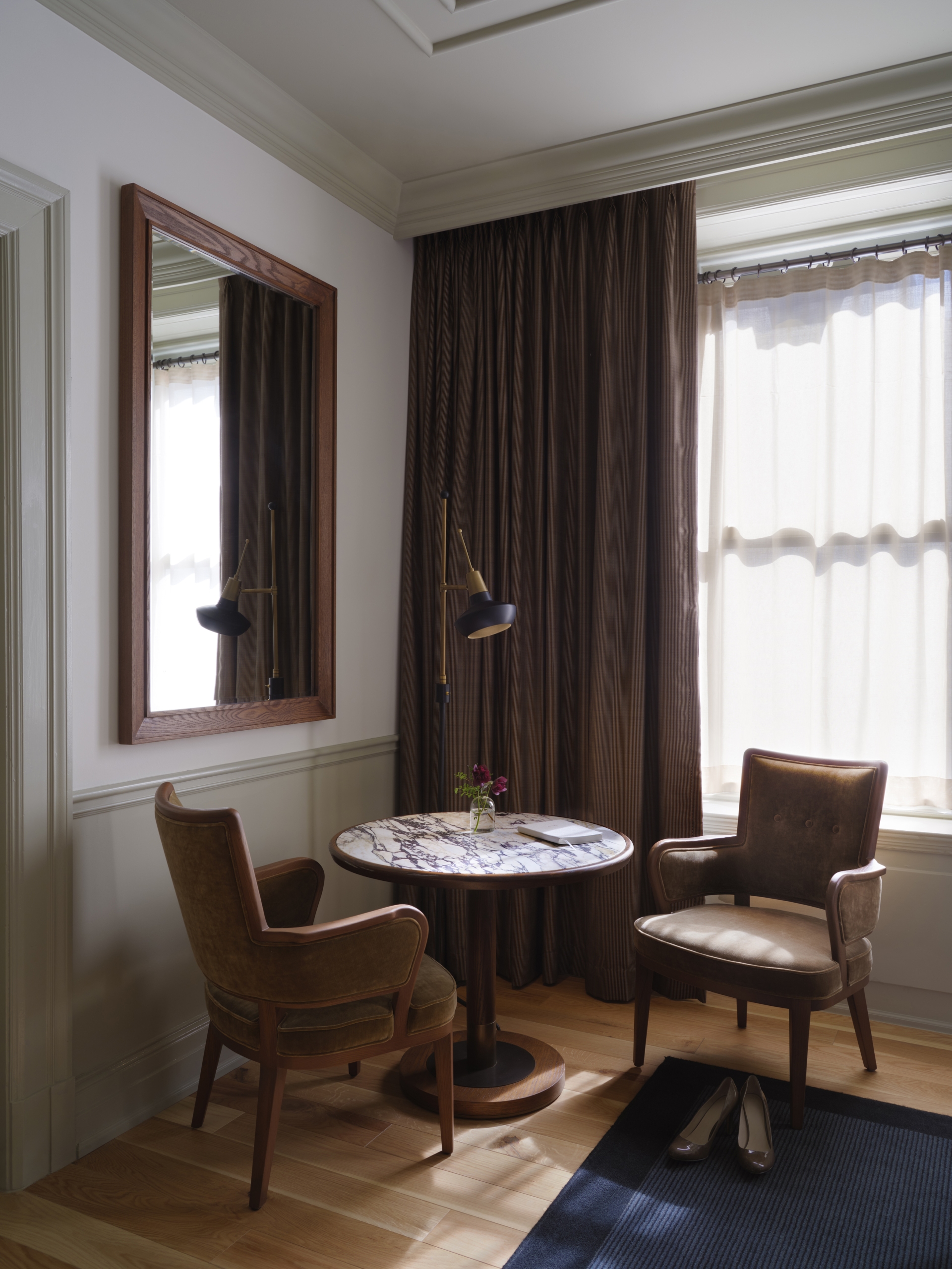 A round marble top table with two slim leather armchairs in front of a window. There is a small flower vase and a floor lamp.