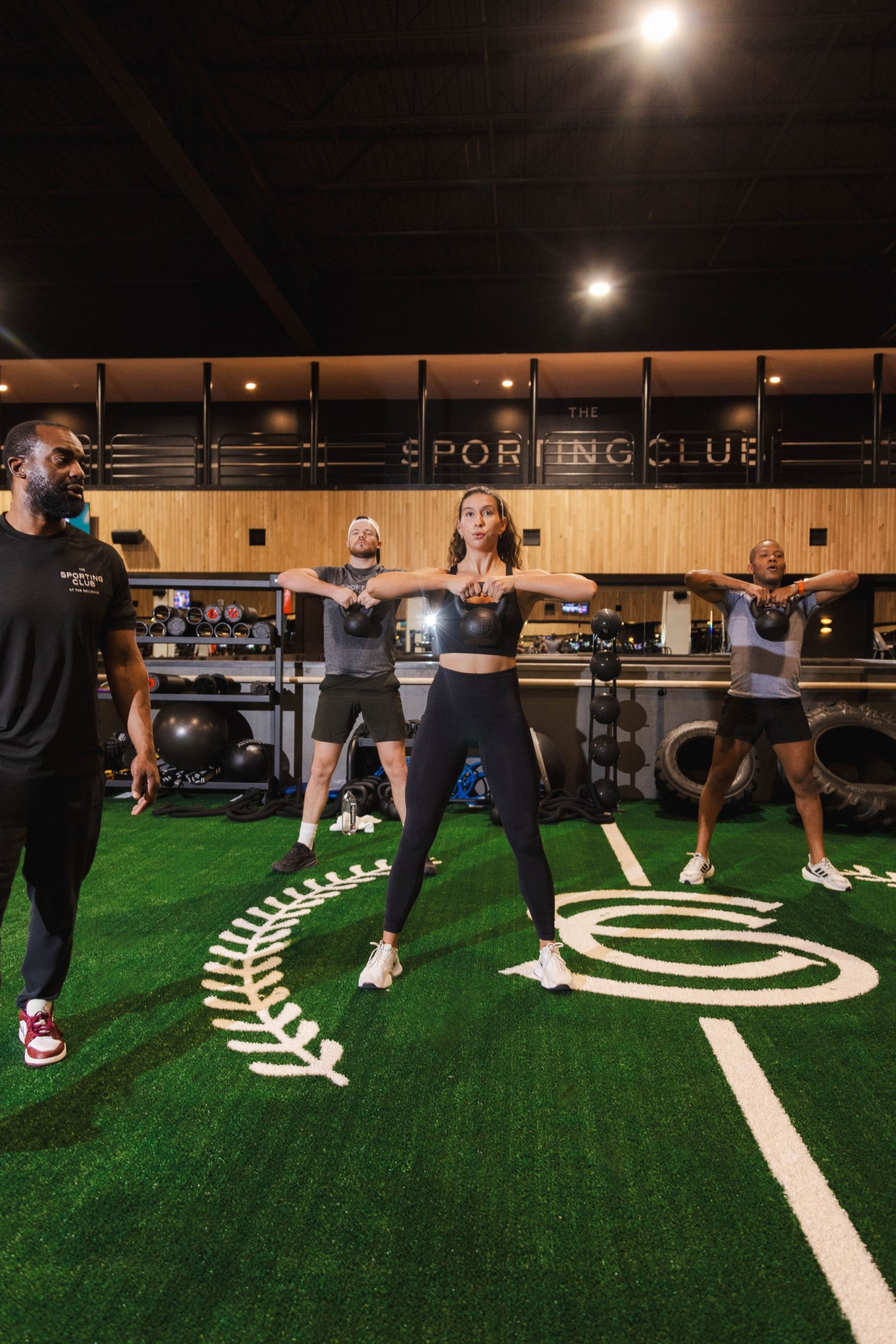 A fitness class with three people lifting kettlebells on a branded turf interior fitness surface. There is an indoor track above and fitness equipment behind