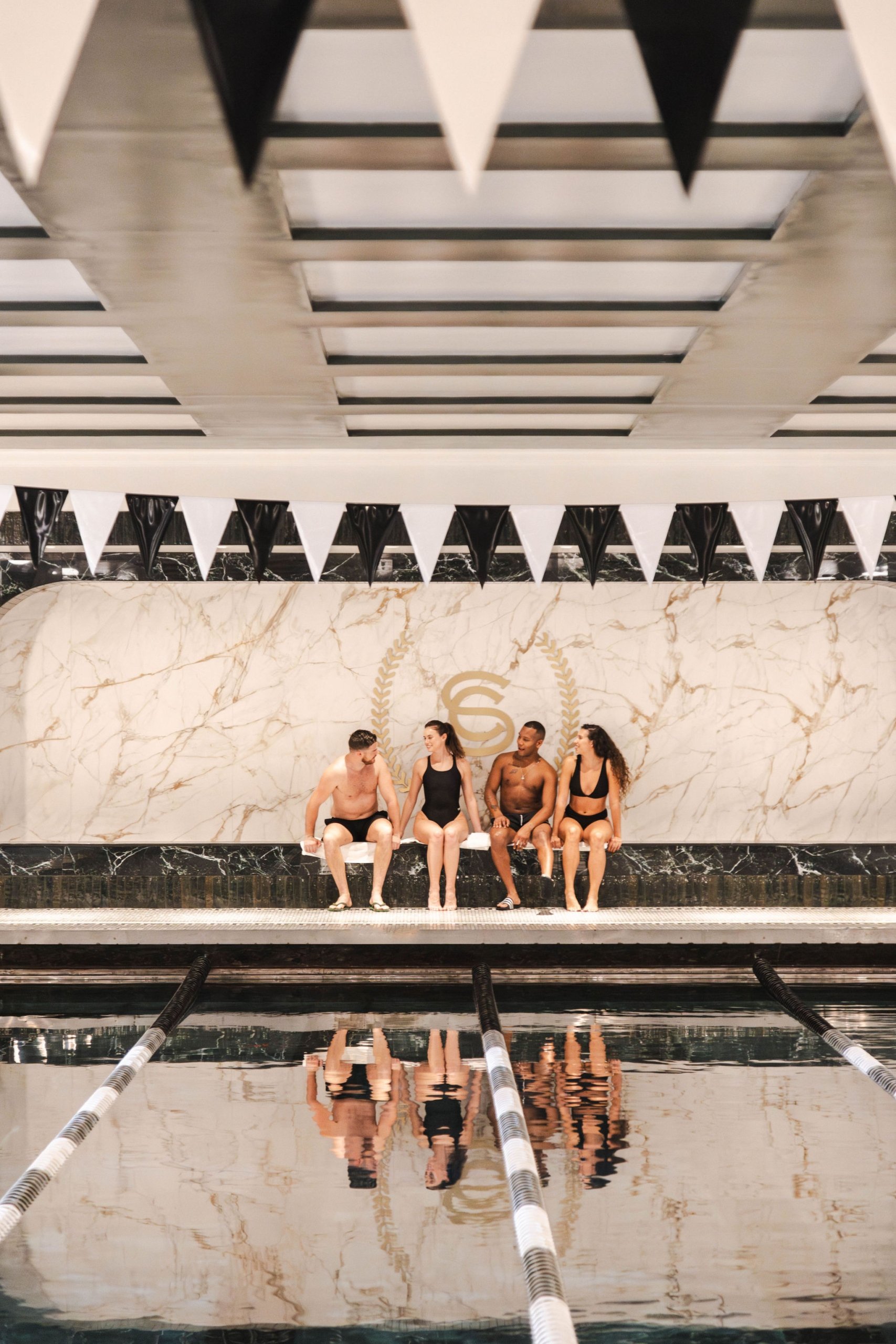 Four friends are sitting in the distance against a marble wall, looking at a reflective lap swimming pool.