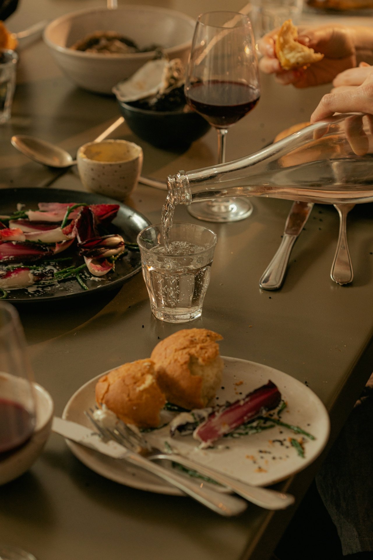 A hand pouring water in a glass with plates full of eaten food.