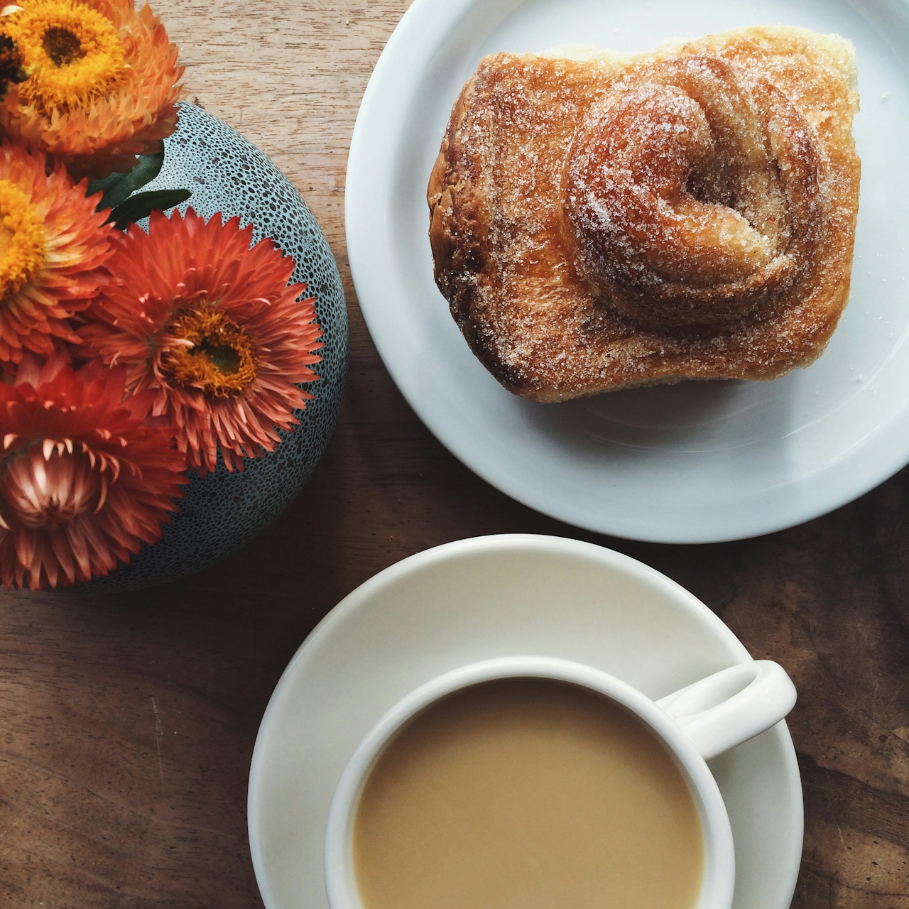 An overhead shot of a cup of coffee and a fresh pastry next to a small bud vase of flowers