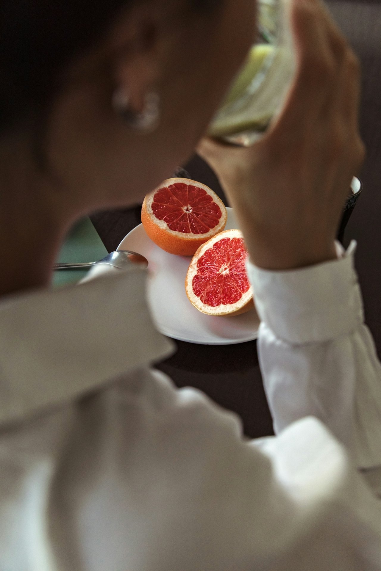 A close over the shoulder shot of a woman drinking from a glass with a plate of grapefruit haves in front of her