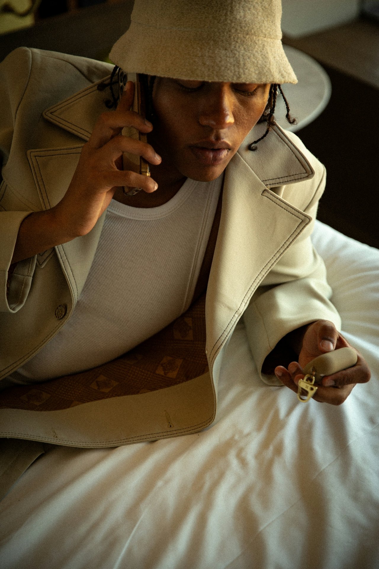 A man in a double breasted jacket and white bucket hat talks on the phone while lying on a white bed