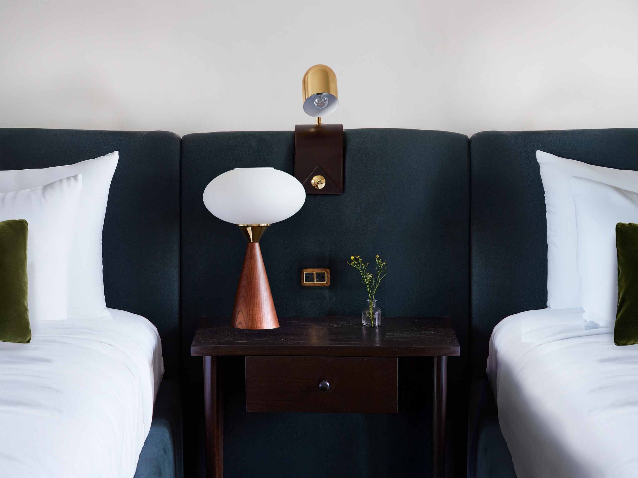 A tight shot of two queen beds with connected navy velvet headboard. There is a mounted reading sconce and a table lamp on the shared side table, and two outlets built into the headboard