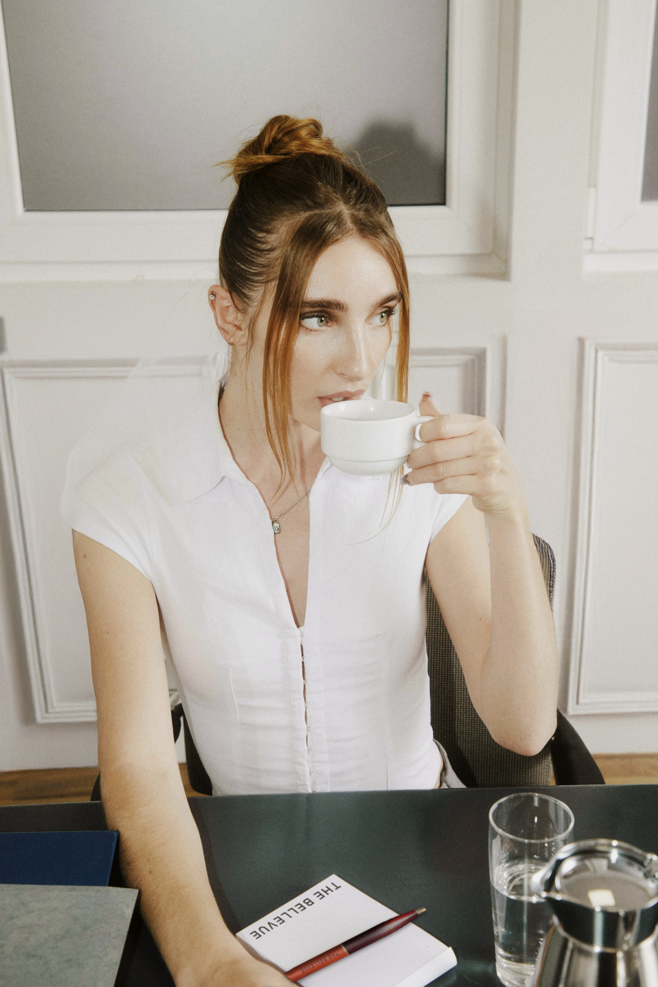 A woman drinking a cup of coffee in business casual attire. There is a branded Bellevue pen and notepad on the desk next to a carafe of water and drinking glass.