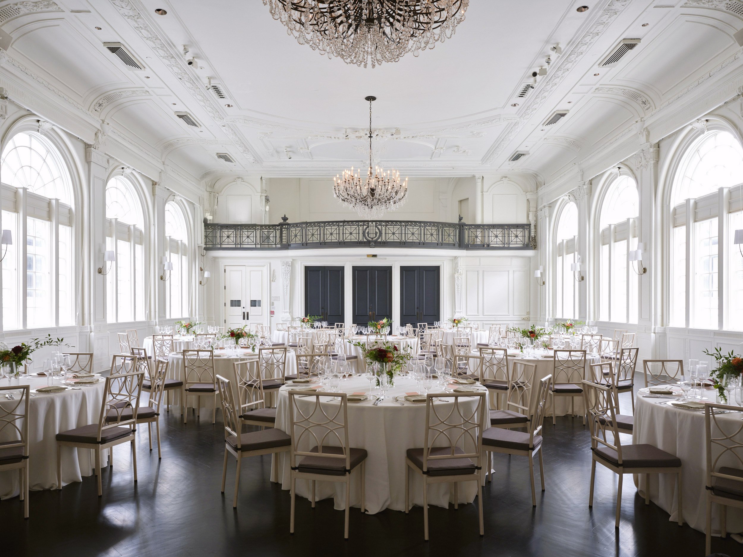 A wide view of a classical white room with high ceilings and ornate moldings. There is a balcony and a chandelier overlooking 10 round tables set for a luncheon