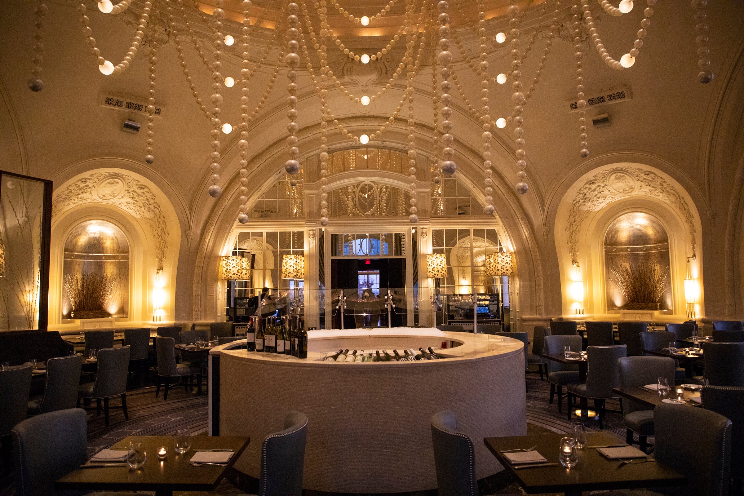 A circular room with a circular bar at the center. there are oversized pearls draped and hanging from the ceiling, and lit coves next to a large arched window