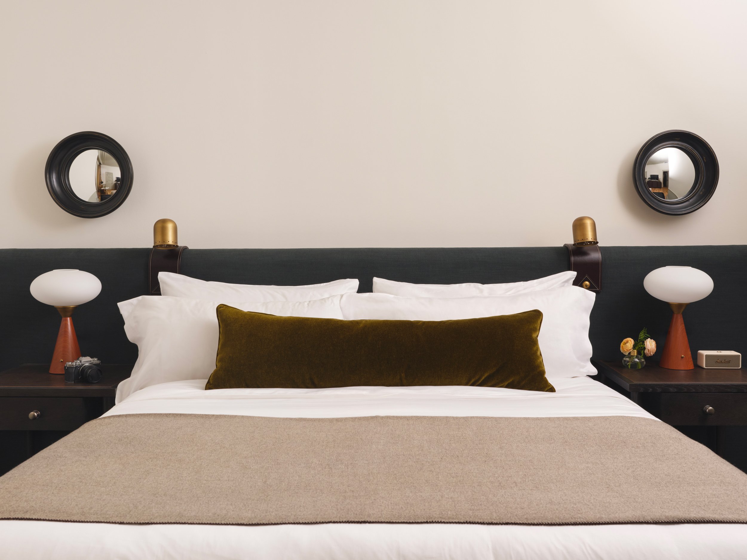 A straight on shot of a king bed headboard, upholstered in navy velvet, with an olive navy bolster pillow. There are matching Mid-Century style table lamps and fisheye mirrors above each nightstand.