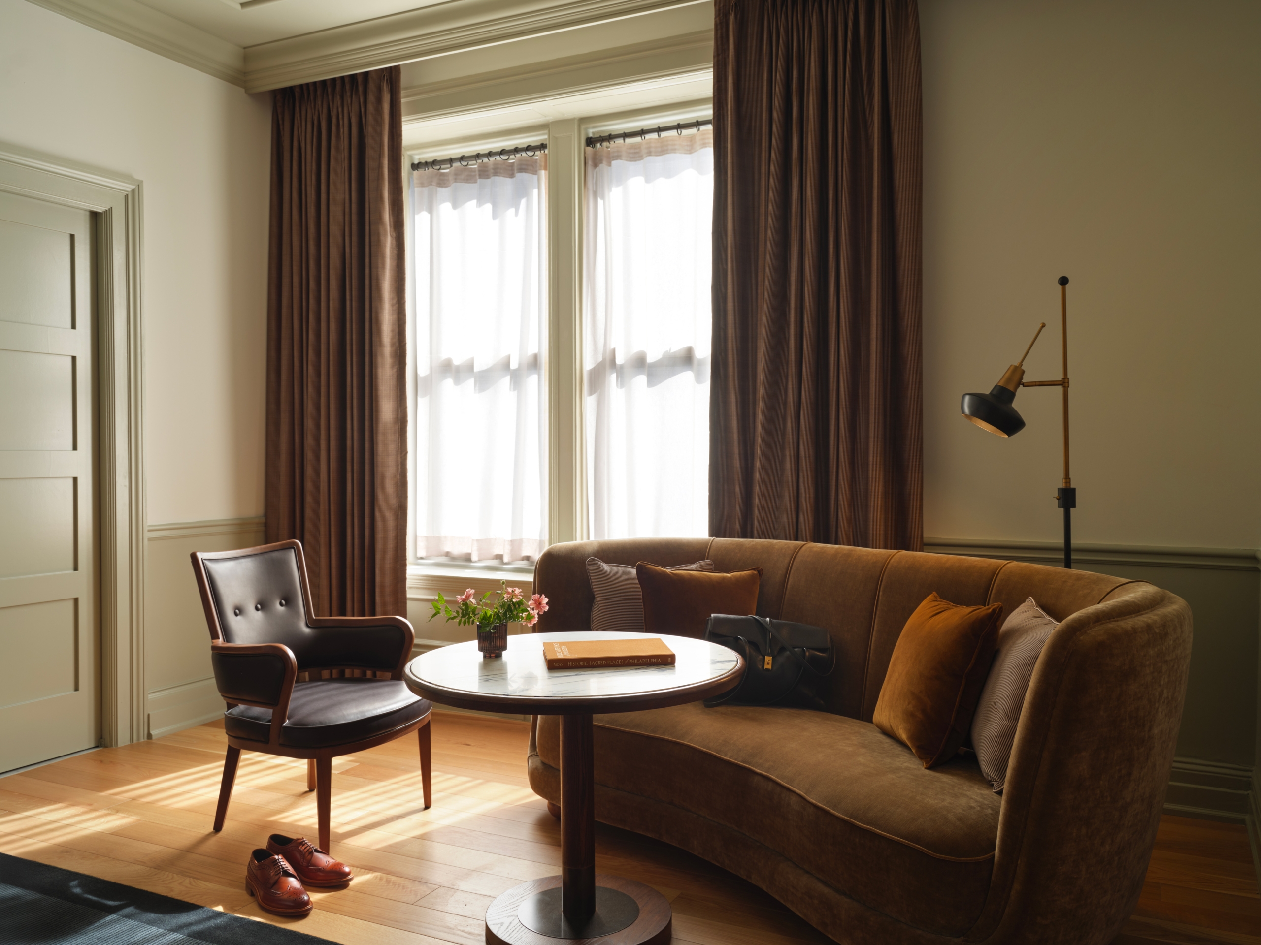 A mustard velvet curved sofa and a slim brown leather armchair face a marble dining height table. There is a book and a floral arrangment on the table, with a black leather bag on the ofa and pair of brown shoes on the floor.