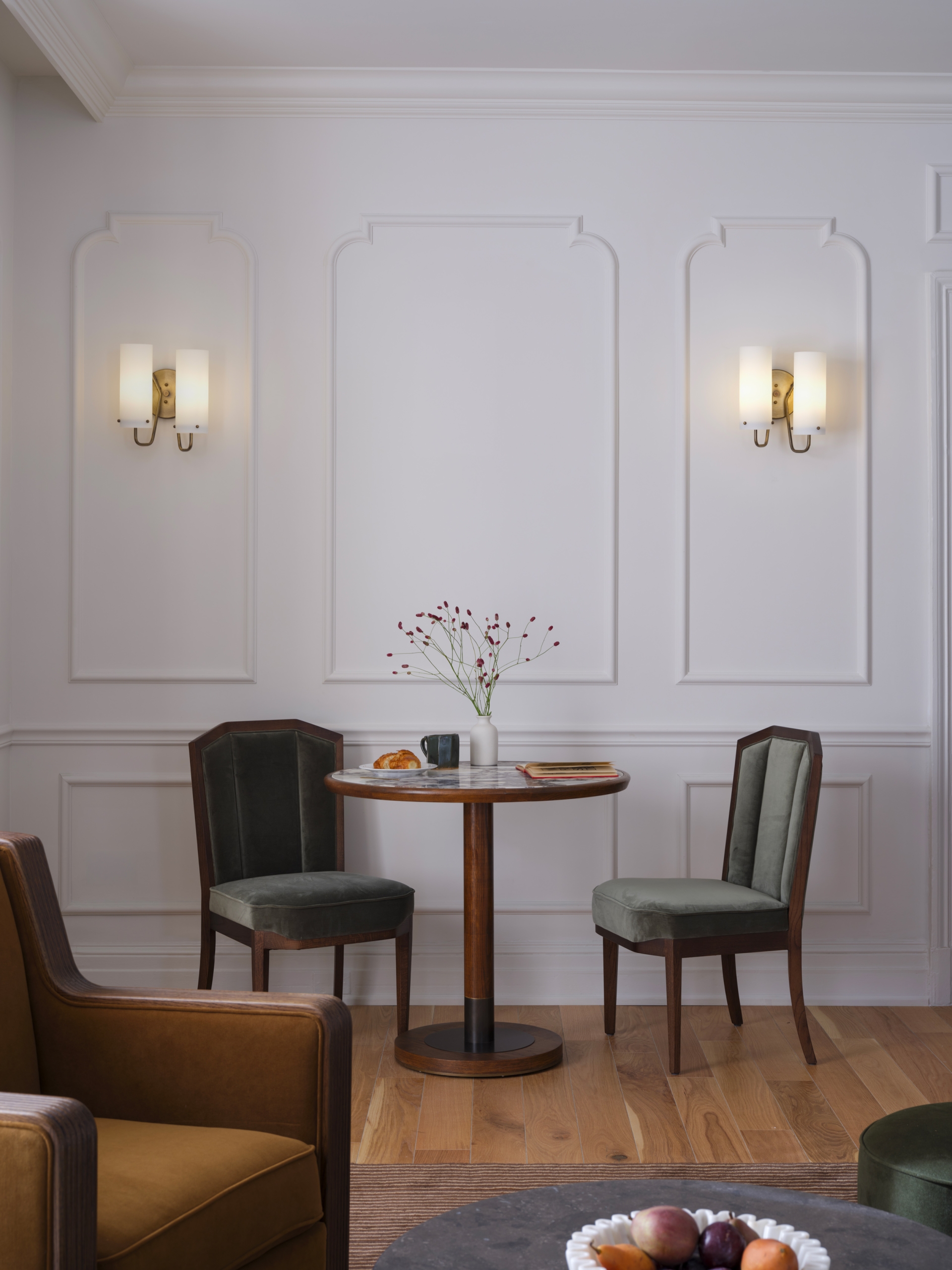 A view of two upholstered dining chairs beside a marble top dining table. There are applied wall moldings and in the foreground a club chair facing a stone coffee table.