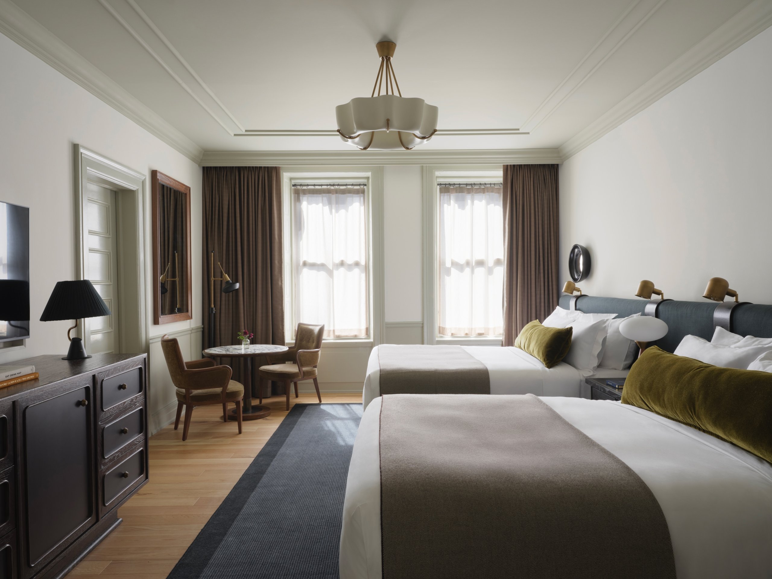 Two queen beds in a gracious room. There is a dark wood dresser with a table lamp, and a two-seat dining table near the window.