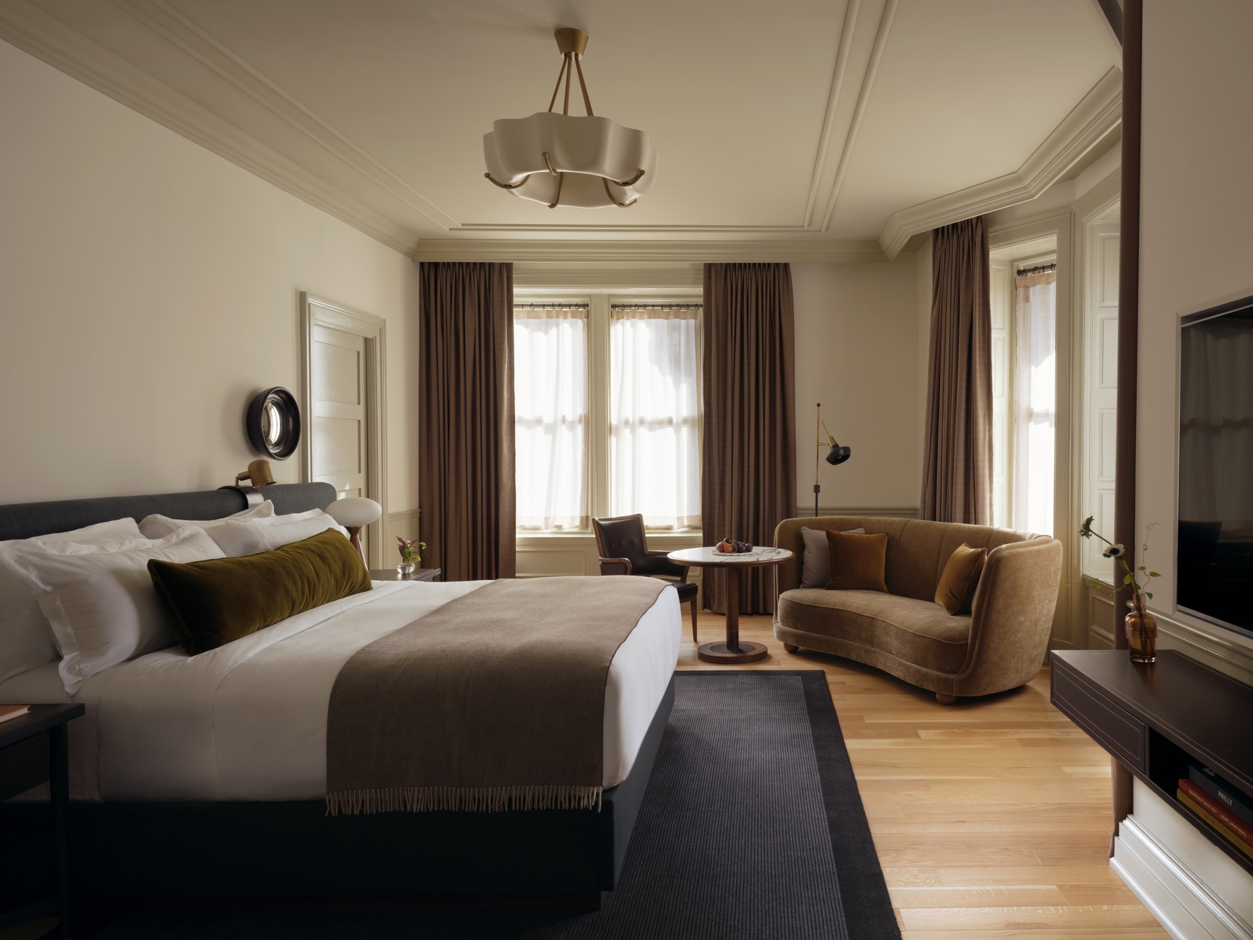 A wide view of a guest room with a velvet upholstered king sized bed. There is a seating area with a velvet sofa and leather chair facing a dining height marble table. A navy carpet lays over wood flooring.